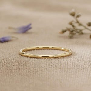 May include: A thin, polished gold band ring with a smooth, rounded surface. The ring features a simple, circular design with a reflective metallic finish that catches the light, showcasing a clean and uniform appearance throughout the entire band.