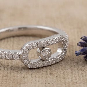 May include: A silver ring with a unique design. The ring features a central bezel-set diamond and is surrounded by a halo of smaller diamonds. The band is also encrusted with small diamonds, creating a sparkling effect. The ring is set against a neutral background.