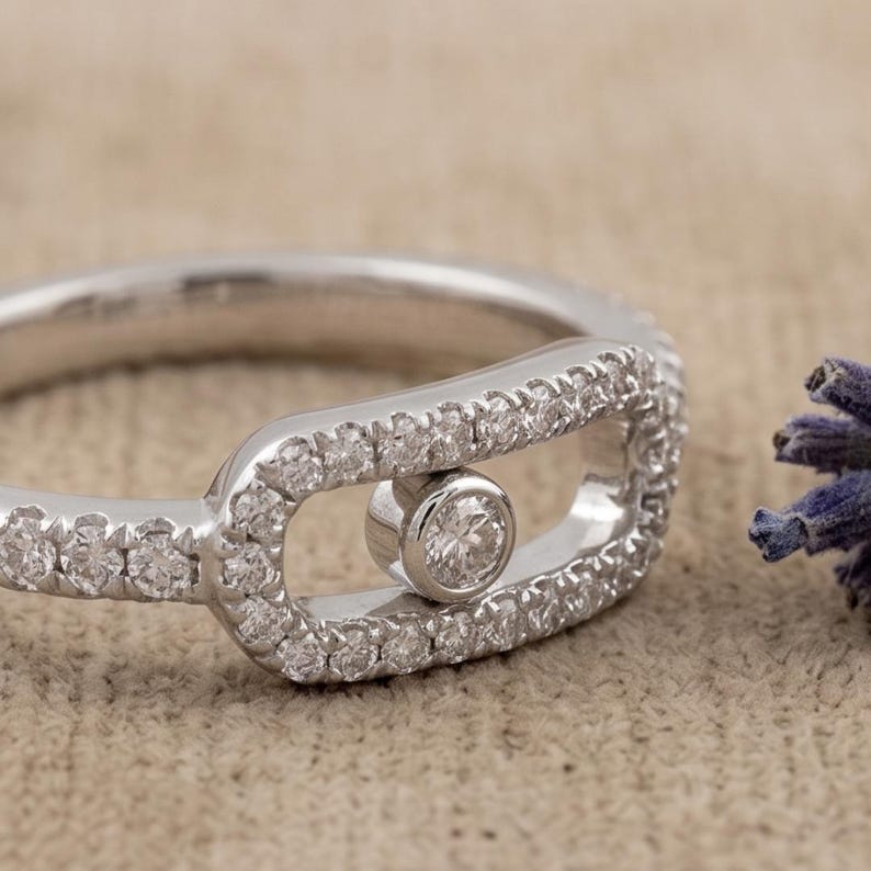 May include: A silver ring with a unique design. The ring features a central bezel-set diamond and is surrounded by a halo of smaller diamonds. The band is also encrusted with small diamonds, creating a sparkling effect. The ring is set against a neutral background.