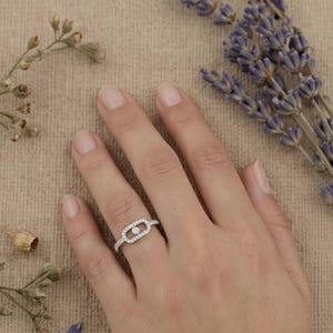 May include: A silver ring with a central oval design, encrusted with small, clear gemstones. A single, larger gemstone is set in the center. The ring is on a finger, with lavender and dried flowers in the background.