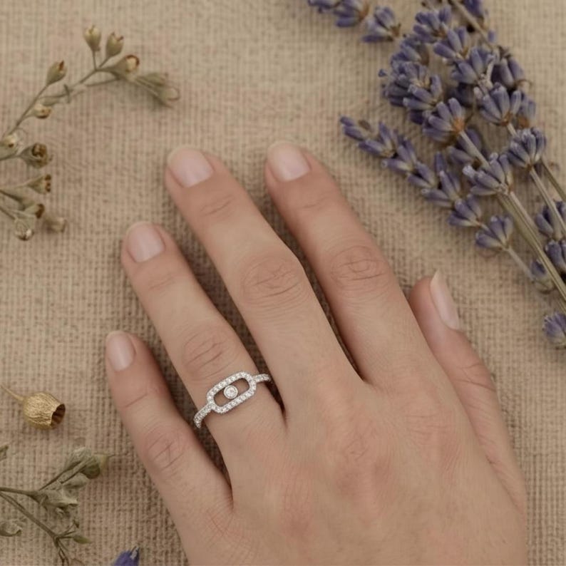 May include: A silver ring with a central oval design, encrusted with small, clear gemstones. A single, larger gemstone is set in the center. The ring is on a finger, with lavender and dried flowers in the background.