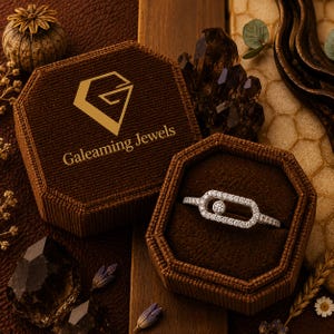 May include: A diamond ring in a brown velvet jewelry box. The ring features a central round diamond surrounded by smaller diamonds. The box is open, revealing the ring, and has the text "Galeaming Jewels" on the lid.