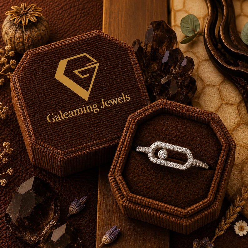 May include: A diamond ring in a brown velvet jewelry box. The ring features a central round diamond surrounded by smaller diamonds. The box is open, revealing the ring, and has the text "Galeaming Jewels" on the lid.