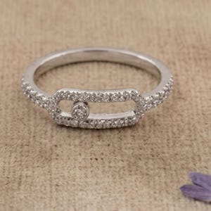 May include: A silver ring with a rectangular center design and a small, round diamond in the middle. The band and rectangular design are encrusted with small, clear stones. The ring is set against a neutral background.