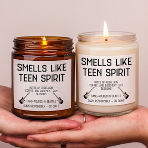 May include: Two lit scented candles in glass jars. The labels read "Smells Like Teen Spirit" with text about rebellion, coffee, and jam sessions. The candles are hand-poured in Seattle. One jar is amber, the other is clear.