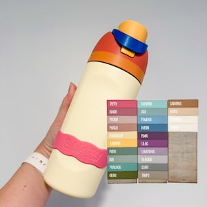May include: A cream-colored water bottle with a pink silicone band featuring the word Oakley, topped with a multi-colored orange, blue, and yellow lid. Beside it is a color chart displaying various shades labeled Taffy, Blush, Peony, Peach, Buttercup, Banana, Fern, Sea, Peacock, Olive, Bahama, Sky, Powder, Denim, Plum, Lilac, Lavender, Glacier, Slate, Taupe, Caramel, Ivory, Coconut, and Sugar.
