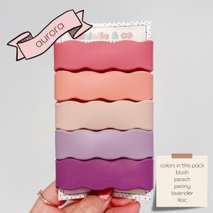 A hand holding a pack of five wavy hair clips in various pastel colors, including pink, peach, peony, lavender, and lilac.