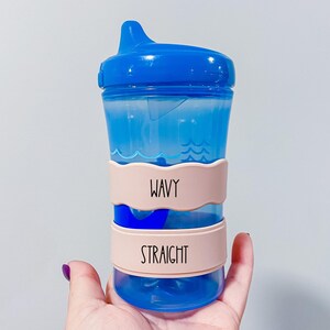 A blue sippy cup with a blue lid and pink straps labeled "Wavy" and "Straight" is being held in a hand.