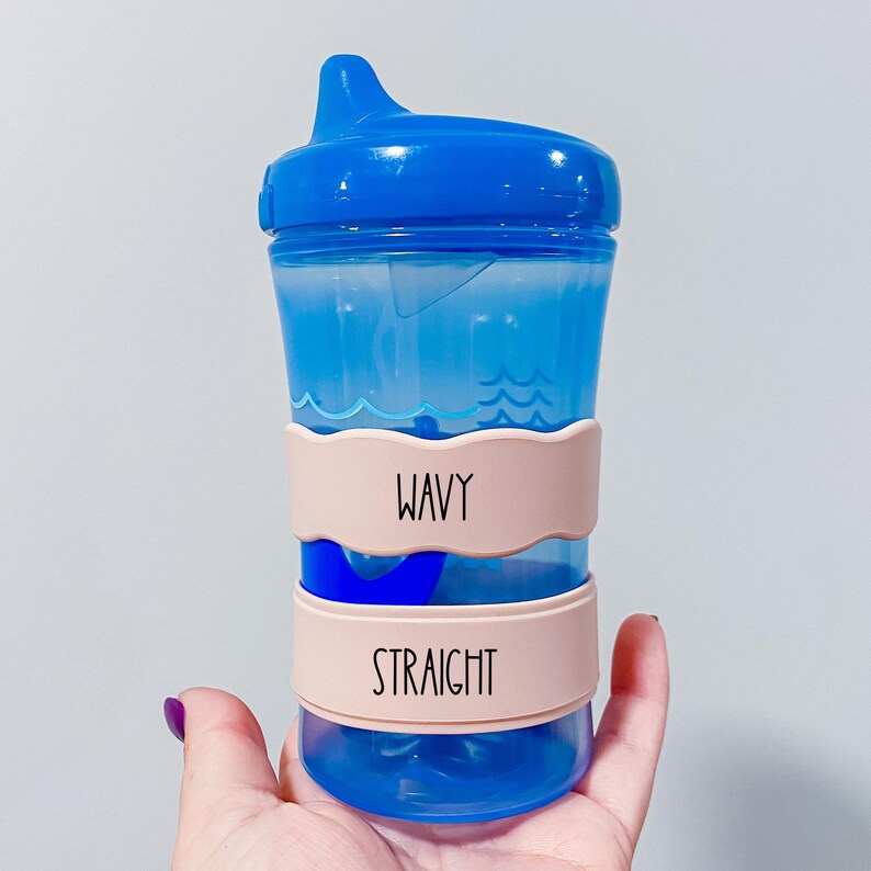 A blue sippy cup with a blue lid and pink straps labeled "Wavy" and "Straight" is being held in a hand.