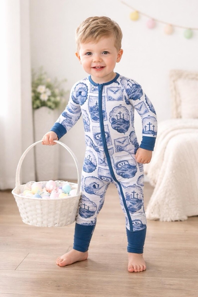 K&ouml;nnte beinhalten: Ein Kleinkind in einem wei&szlig;-blau gemusterten Strampler mit Rei&szlig;verschluss h&auml;lt einen wei&szlig;en Weidenkorb mit pastellfarbenen Ostereiern. Der Strampler hat einen blauen Aufdruck mit Kreuzen und anderen Designs.