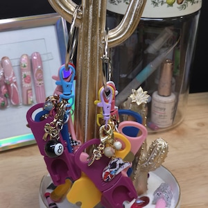 Könnte beinhalten: Eine Sammlung bunter Flaschenöffner aus Kunststoff an silbernen Metallschlüsselringen, die an einem goldfarbenen kaktusförmigen Ständer hängen. Die Öffner sind in Lila, Gelb und Pink gehalten und mit kleinen Anhängern wie einer Achtkugel versehen.
