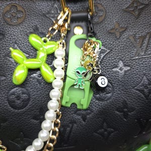 Può includere: Una borsa in pelle nera con motivo monogramma presenta un set di ciondoli decorativi. I ciondoli includono un cane a palloncino verde lime, una fila di perle bianche, una clip in plastica verde, una figura aliena, un fiocco nero e una palla numero 8.