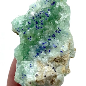 Puede incluir: Primer plano de un espécimen mineral con una estructura cristalina verde claro y acentos azul oscuro. El mineral tiene una superficie rugosa y texturizada y una base beige. La apariencia general es natural y orgánica.