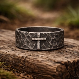 以下が含まれることがあります： ハンマー加工が施された、十字架のデザインが特徴の、テクスチャ加工された濃いグレーのリング。木製の表面に置かれ、素朴な美しさを際立たせています。リングの幅は約1.3cmです。
