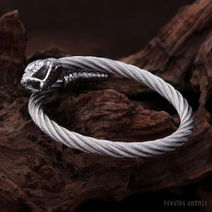 May include: A silver-toned metal bracelet constructed from twisted wire cable. The bracelet features a detailed snake head at one end and a tapered tail at the other, forming an open-ended circular design. The text Forging Gnomes is visible.