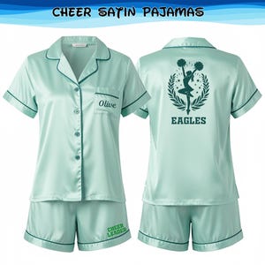 May include: A mint green satin pajama set, comprising a button-down shirt and shorts. The shirt features the name "Olive" embroidered on the pocket. The shorts are printed with "Cheer Leader". The shirt's back displays a cheerleader graphic and the word "EAGLES".