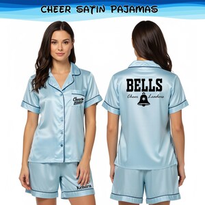 May include: Light blue satin pajama set with black trim. The short-sleeved top has a button-down front and the text "Cheer Leader" on the front. The shorts have the name "Kendra" embroidered on the leg. The back of the top says "BELLS Cheer Leaders".