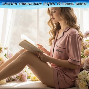 May include: Pink rayon pajama set with black trim, featuring the word "Bride" embroidered on the pocket. The set includes a short-sleeved top and shorts. The image also includes the text "CUSTOM EMBROIDERY RAYON PAJAMAS SHORT".