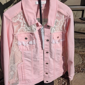 May include: A light pink denim jacket with white lace panels on the front and sleeves. The jacket has a button-up closure, two front pockets, and a classic collar. The lace detailing adds a feminine touch to the casual style.