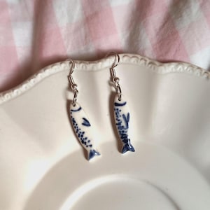 Op de afbeelding: Een paar keramische visoorbellen. De oorbellen zijn wit met blauwe details, waaronder stippen en vinnen. Ze hangen aan zilverkleurige haken. De oorbellen worden gepresenteerd op een wit bord.