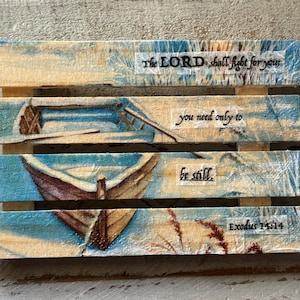 May include: Wooden art piece with four horizontal planks, featuring a boat scene and inspirational text. The top plank reads "The LORD shall fight for you." Other planks include "you need only to" and "be still." The bottom plank reads "Exodus 14:14."