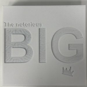 Könnte beinhalten: Quadratische weiße Oberfläche mit den Worten "The Notorious BIG" in erhabener, strukturierter Schrift. Die Buchstaben sind mit einem Holzmaserungsmuster gefüllt. Eine kleine Krone befindet sich in der unteren rechten Ecke.