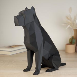 Könnte beinhalten: Eine mattschwarze Hundefigur in geometrischem Low-Poly-Design. Die Skulptur besteht aus scharfen, kantigen Facetten, die den Kopf, die Ohren und den Körper eines sitzenden Hundes bilden, und ist durchgehend in einem dunklen, einfarbigen Ton gehalten.