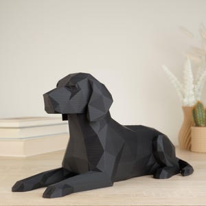 Może przedstawiać: Czarna figurka psa wykonana w geometrycznym stylu low-poly. Rzeźba przedstawia psa w pozycji leżącej z uniesioną głową, charakteryzującą się ostrymi, kanciastymi płaszczyznami, które precyzyjnie definiują jego formę oraz całą strukturę.