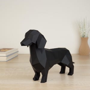 Könnte beinhalten: Eine schwarze, geometrische Dackel-Skulptur. Die Hundefigur besteht aus eckigen Formen, was ihr einen modernen, minimalistischen Look verleiht. Die Skulptur steht auf einer hellen Holzoberfläche. Ein Stapel Bücher und eine Vase mit Blumen befinden sich im Hintergrund.