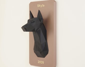 Puede incluir: Una escultura de cabeza de perro geométrica negra de bajo polígono montada sobre una placa rectangular de color marrón claro. El nombre Skyla está impreso en texto claro sobre la cabeza del perro y el número 2026 debajo de ella.