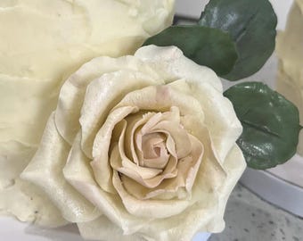 May include: Close-up of a cake decorated with a large, cream-colored rose and green leaves. The rose has a delicate, layered petal design with a soft, light brown center. The cake has a textured, off-white frosting.