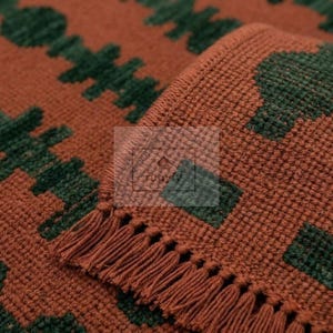 Peut inclure: Un textile tissé présentant une base de couleur rouille avec un motif géométrique vert foncé répétitif. Le tissu a une surface texturée et granuleuse, avec un bord fini par des franges courtes et denses. Un logo rectangulaire avec le texte TUUVA est visible.
