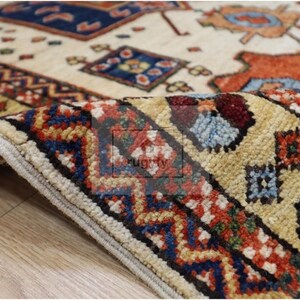 Puede incluir: Primer plano de una alfombra estampada enrollada, con un fondo beige y diseños intrincados en rojo, azul y naranja. El borde de la alfombra presenta un patrón geométrico detallado. La alfombra está sobre una superficie de madera.