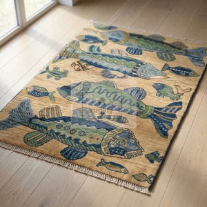 Op de afbeelding: Een rechthoekig vloerkleed met een beige achtergrond en een herhalend patroon van gestileerde vissen in blauwe, groene en bruine tinten. Het kleed heeft franjes aan de boven- en onderkant en bevat de tekst rugviy in het midden.