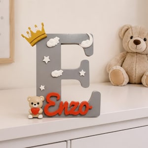 Op de afbeelding: Een grijze houten letter "E" versierd met witte wolken, sterren en een gouden kroon. De naam "Enzo" is in het rood geschreven. Een klein teddybeerbeeldje met een rood hart staat naast de letter. Een knuffelbeer staat op de achtergrond.