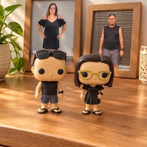Puede incluir: Dos figuras coleccionables de vinilo de pie una al lado de la otra. La figura de la izquierda tiene el pelo castaño corto, gafas de sol negras, una camiseta negra sin mangas, pantalones cortos grises y chanclas negras. La de la derecha tiene el pelo oscuro, gafas amarillas, un vestido negro y chanclas negras.