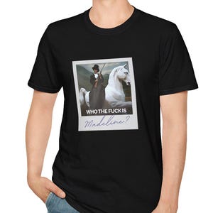 Op de afbeelding: Zwart t-shirt met een afbeelding van een persoon op een wit paard, met de tekst "WHO THE FUCK IS Madeline?" op een fotolijst in polaroidstijl. Het shirt is gemaakt van een zacht materiaal en wordt door een persoon gedragen.