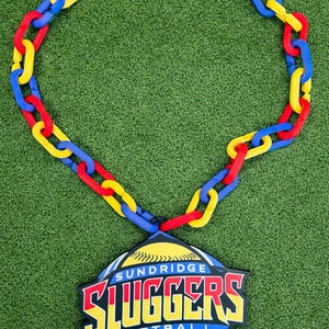 Peut inclure: Un grand collier composé d'une chaîne de maillons en plastique rouge, jaune et bleu. Il est doté d'un pendentif en forme de losange portant l'inscription "SUNDRIDGE SLUGGERS SOFTBALL MVP" en lettres jaunes, rouges et blanches sur fond bleu.