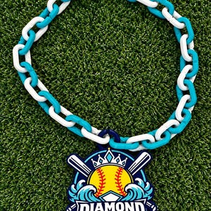 Peut inclure: Un collier composé d'une chaîne à maillons en plastique alternant le blanc et le turquoise. Un pendentif est suspendu à la chaîne, représentant une balle de softball jaune à coutures rouges, deux battes de baseball blanches croisées, une couronne et des vagues bleues avec le texte Diamond Queen.