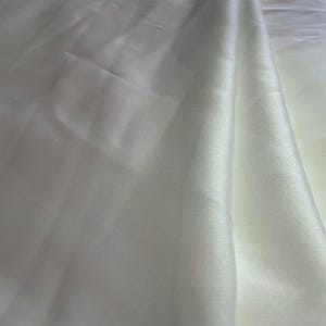 K&ouml;nnte beinhalten: Nahaufnahme eines wei&szlig;en, seidigen Stoffes mit weichen Falten. Das Material hat einen subtilen Glanz, der das Licht auf seiner Oberfl&auml;che reflektiert. Der Stoff wirkt glatt und flie&szlig;end, was auf ein luxuri&ouml;ses Textil hindeutet.