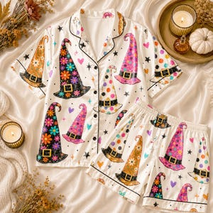 May include: A white short-sleeve button-down pajama shirt and matching shorts set featuring a colorful pattern of witch hats decorated with flowers and polka dots, interspersed with small black stars and pink, orange, and blue hearts.