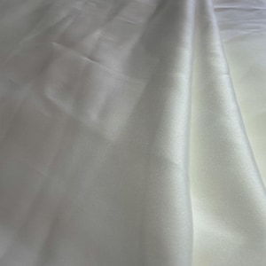 May include: A length of solid white fabric with a smooth, lustrous surface and a subtle sheen. The material is draped in soft, flowing folds, creating gentle shadows and highlights across the surface that emphasize its reflective quality.