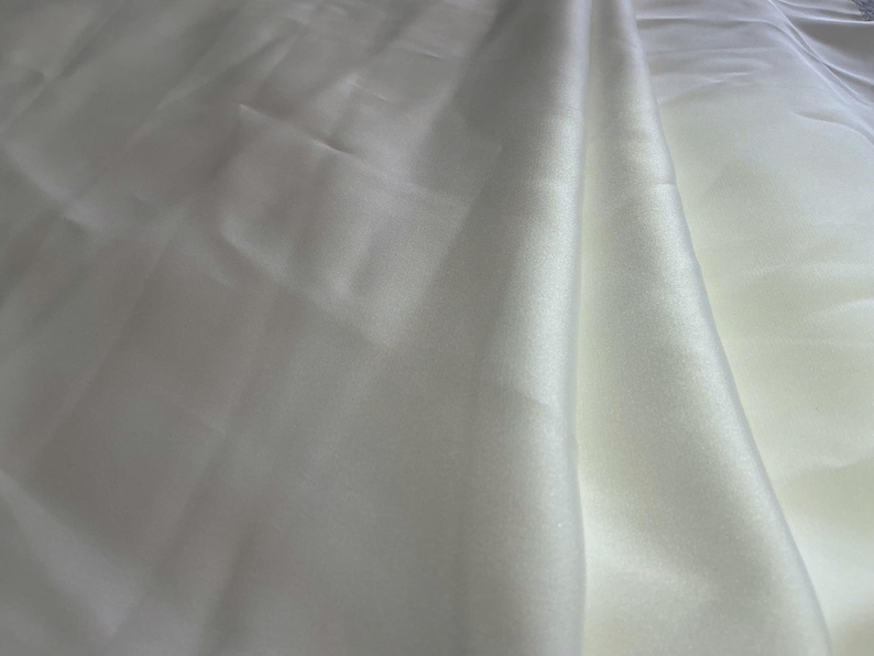 May include: A length of solid white fabric with a smooth, lustrous surface and a subtle sheen. The material is draped in soft, flowing folds, creating gentle shadows and highlights across the surface that emphasize its reflective quality.