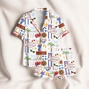 May include: White casino-themed pajama set featuring a short-sleeved button-up shirt and matching shorts. The set is adorned with casino-related graphics, including dice, playing cards, and the text "Welcome Las Vegas". The trim is red.