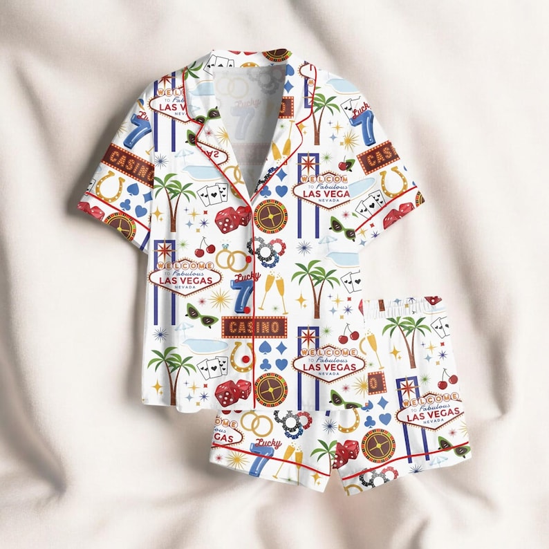 May include: White casino-themed pajama set featuring a short-sleeved button-up shirt and matching shorts. The set is adorned with casino-related graphics, including dice, playing cards, and the text "Welcome Las Vegas". The trim is red.