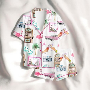 Puede incluir: Conjunto de pijama blanco con un estampado vibrante que presenta flamencos rosas, coches antiguos, m&aacute;quinas tragamonedas, dados y copas de c&oacute;ctel. El conjunto incluye una camisa con botones y pantalones cortos a juego, ribeteados con ribetes rosas.