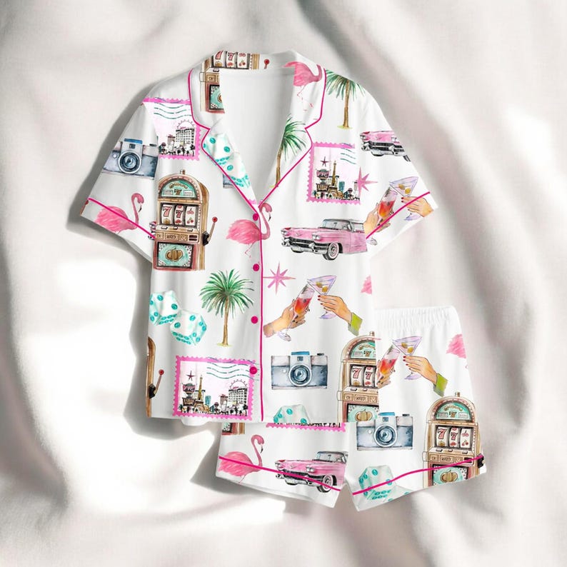 Puede incluir: Conjunto de pijama blanco con un estampado vibrante que presenta flamencos rosas, coches antiguos, m&aacute;quinas tragamonedas, dados y copas de c&oacute;ctel. El conjunto incluye una camisa con botones y pantalones cortos a juego, ribeteados con ribetes rosas.
