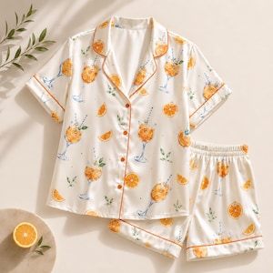 May include: A two-piece pajama set featuring a short-sleeve button-down shirt and matching shorts. The fabric is cream-colored with an all-over print of orange slices and glasses of orange juice with straws, accented by orange piping along the edges.