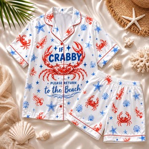 May include: A white short-sleeve button-down pajama shirt and matching shorts set featuring a red crab graphic, blue starfish, and seashell patterns. The shirt includes the text IF CRABBY PLEASE RETURN to the Beach in blue lettering.
