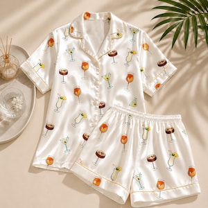 May include: A white short-sleeve button-down pajama shirt and matching shorts set featuring a repeating pattern of various colorful cocktails in glasses with straws and fruit garnishes, finished with thin gold piping along the edges.